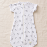 A white short-sleeve baby sleep sack with a front zipper, featuring a repeating pattern of small black line-drawn cartoon faces, laid flat on a beige fabric background.