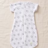 A white short-sleeve baby sleep sack with a front zipper, featuring a repeating pattern of small black line-drawn cartoon faces, laid flat on a beige fabric background.