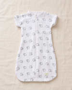 A white short-sleeve baby sleep sack with a front zipper, featuring a repeating pattern of small black line-drawn cartoon faces, laid flat on a beige fabric background.