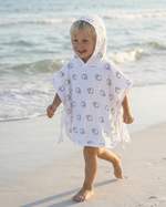 A young child walking barefoot on a sandy beach wearing a white hooded poncho with a black cartoon animal print.