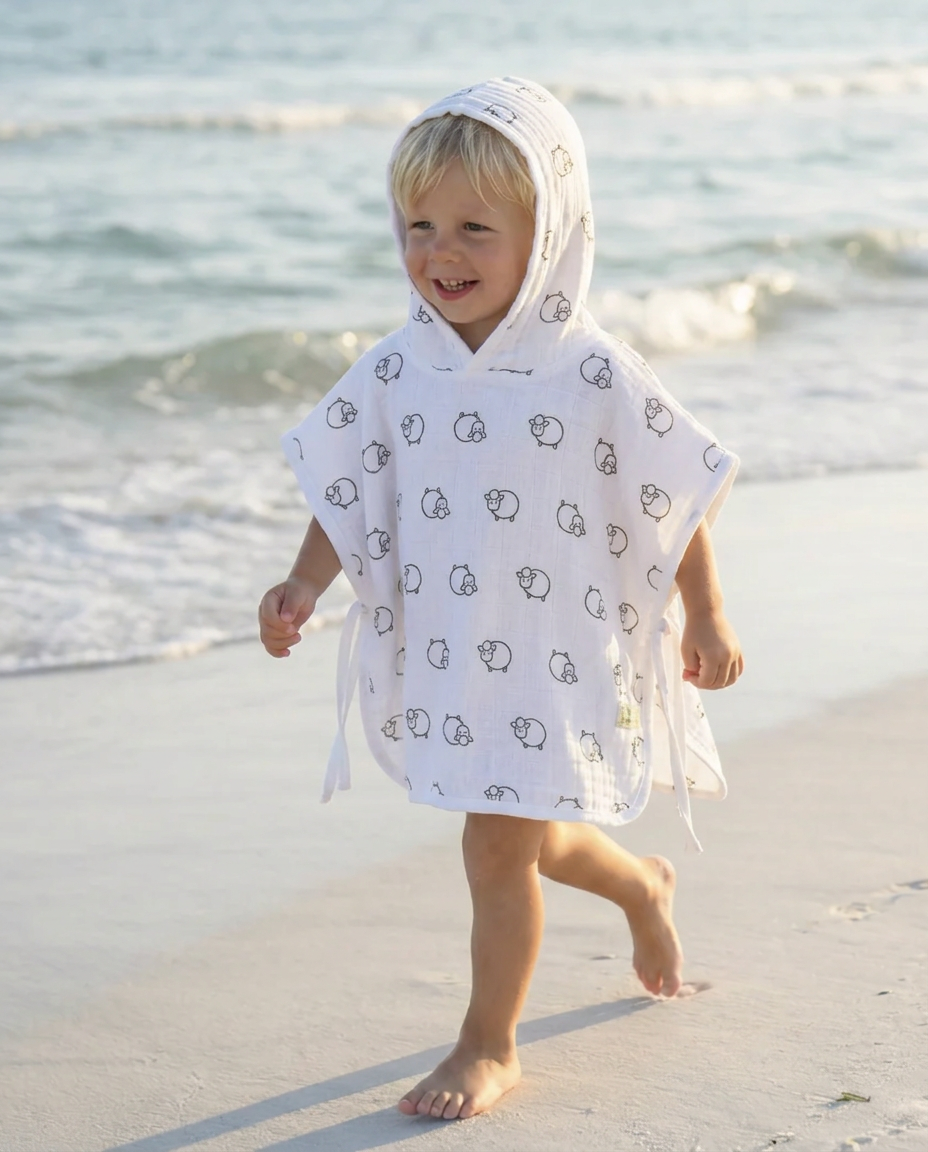 A young child walking barefoot on a sandy beach wearing a white hooded poncho with a black cartoon animal print.