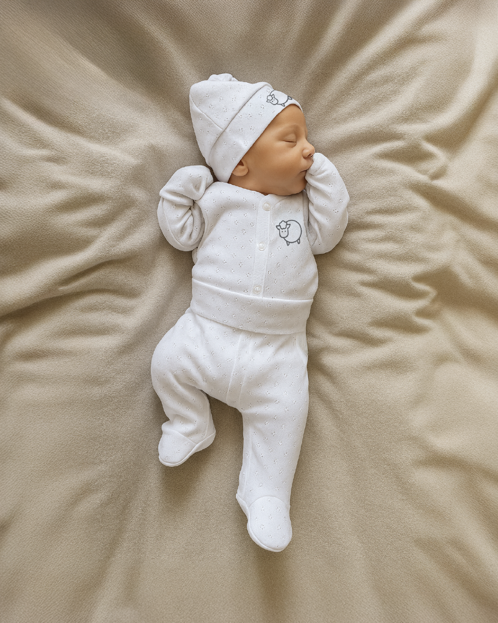 Newborn baby in a white outfit and hat lying on a beige blanket