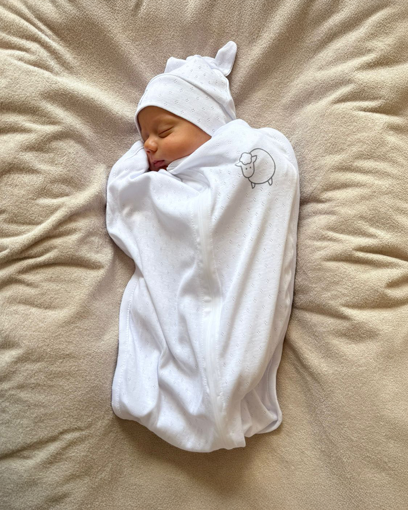 Newborn baby swaddled in white blanket with hat on beige surface