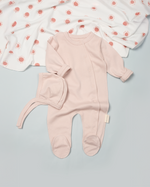 Pink baby onesie with hat on a light gray surface with a white blanket with red patterns in the background.
