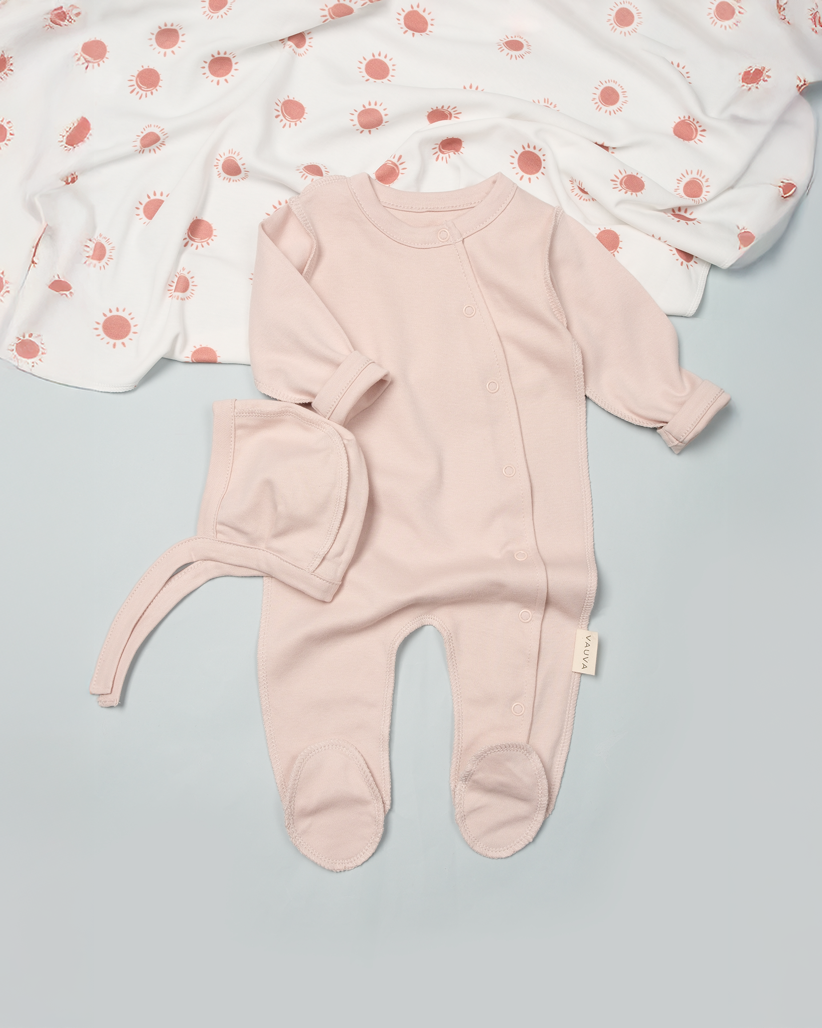 Pink baby onesie with hat on a light gray surface with a white blanket with red patterns in the background.