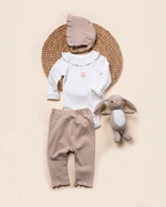 Baby outfit with white top, brown pants, and hat, accompanied by a plush toy on a beige background.