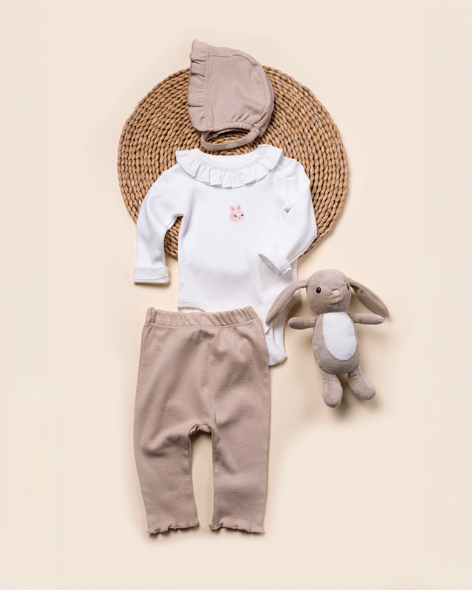 Baby outfit with white top, brown pants, and hat, accompanied by a plush toy on a beige background.