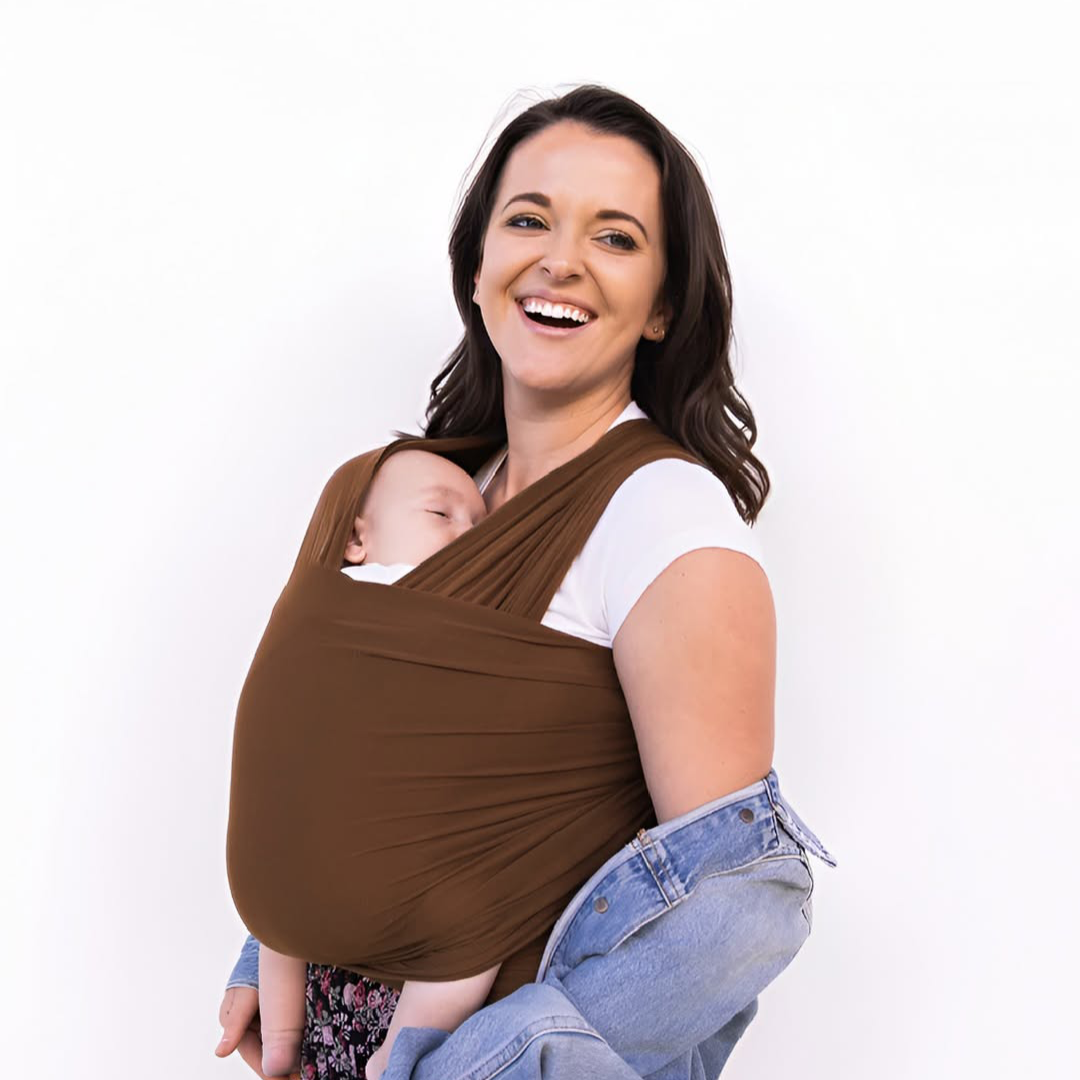 A smiling person is holding an infant in a brown baby wrap carrier.