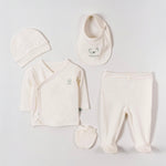 A baby clothing set laid out on a light gray background, including a cream-colored wrap shirt, matching pants, a bib, a beanie, and a small bootie. The bib features a small teddy bear graphic.